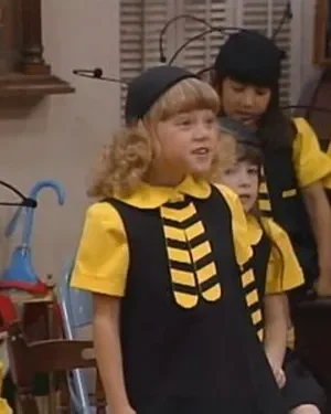 Crianças em fantasias de abelha (pretas e amarelas, chapéus com antenas) em um ambiente interno. Uma menina loira sorri no centro.