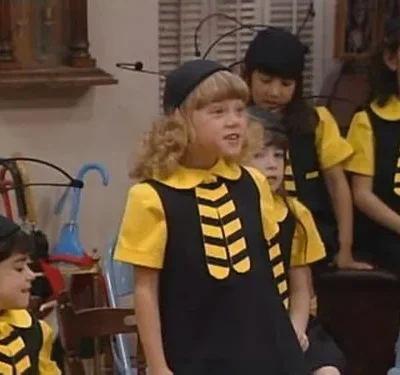 Crianças em fantasias de abelha (pretas e amarelas, chapéus com antenas) em um ambiente interno. Uma menina loira sorri no centro.