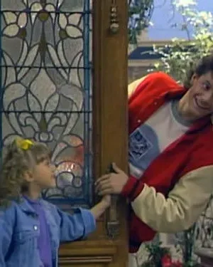 Garota de cabelo cacheado e jaqueta jeans sorri para homem de jaqueta colegial, espiando pela porta. Clima alegre e familiar.