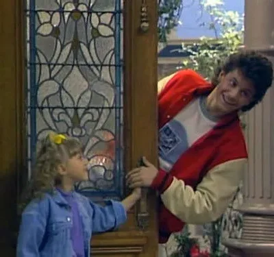 Garota de cabelo cacheado e jaqueta jeans sorri para homem de jaqueta colegial, espiando pela porta. Clima alegre e familiar.