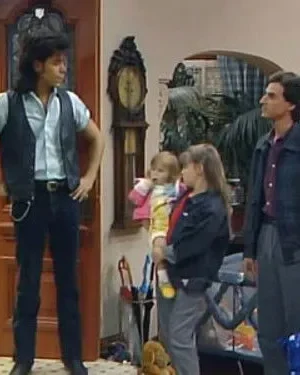 Um homem de colete e outro de jaqueta com duas meninas em casa. Ambiente familiar e descontraído.