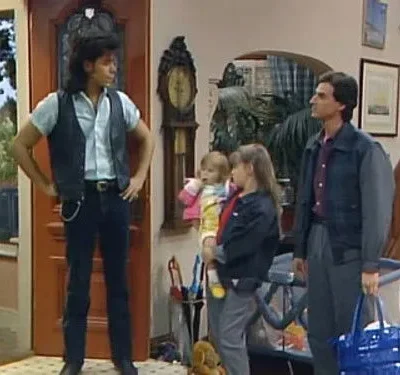 Um homem de colete e outro de jaqueta com duas meninas em casa. Ambiente familiar e descontraído.