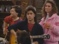 Um homem de mullet toca violão sério, outro sorri ao violão. Uma mulher de rosa com caneca observa. Fundo rústico. Clima musical.