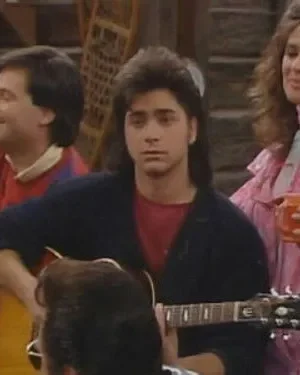 Um homem de mullet toca violão sério, outro sorri ao violão. Uma mulher de rosa com caneca observa. Fundo rústico. Clima musical.