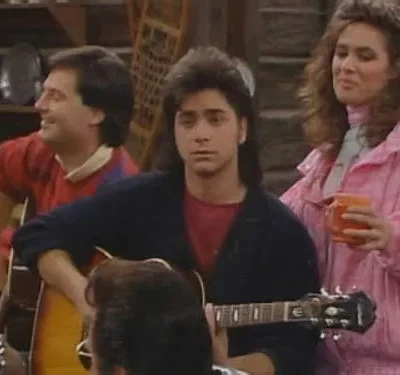 Um homem de mullet toca violão sério, outro sorri ao violão. Uma mulher de rosa com caneca observa. Fundo rústico. Clima musical.