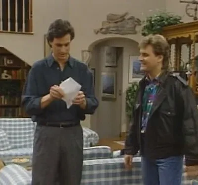 Dois homens numa sala. Um lê um documento com semblante sério, enquanto o outro, de jaqueta, observa sorrindo. Clima doméstico.