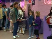 Homem com bebê e duas meninas em loja vibrante dos anos 80/90. Paredes de tijolo rosa, letreiros neon e revistas de música.