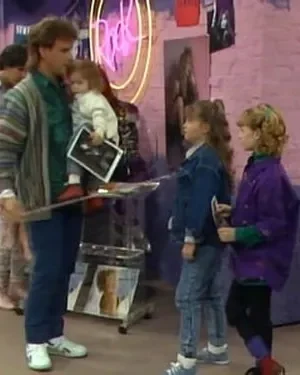 Homem com bebê e duas meninas em loja vibrante dos anos 80/90. Paredes de tijolo rosa, letreiros neon e revistas de música.