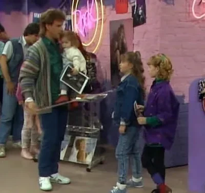 Homem com bebê e duas meninas em loja vibrante dos anos 80/90. Paredes de tijolo rosa, letreiros neon e revistas de música.