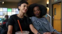 Duas mulheres em um lounge. Uma sorri, de cabelo curto. A outra, de cabelo afro e batom roxo, veste blusa rasgada. Cena casual.