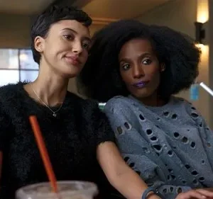 Duas mulheres em um lounge. Uma sorri, de cabelo curto. A outra, de cabelo afro e batom roxo, veste blusa rasgada. Cena casual.