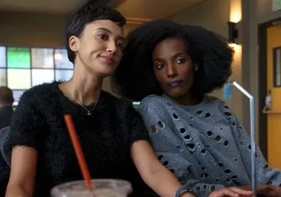 Duas mulheres em um lounge. Uma sorri, de cabelo curto. A outra, de cabelo afro e batom roxo, veste blusa rasgada. Cena casual.