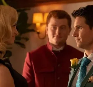 Mulher loira e homem de terno verde sorriem um para o outro. Um homem de uniforme vermelho sorri ao fundo. Cena interna, clima formal.