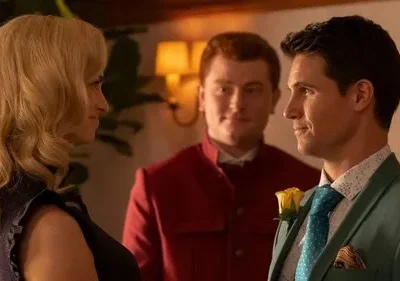 Mulher loira e homem de terno verde sorriem um para o outro. Um homem de uniforme vermelho sorri ao fundo. Cena interna, clima formal.
