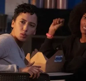 Duas mulheres em ambiente interno. Uma com cabelo encaracolado tem expressão séria. A outra, com afro, sorri e mostra o punho.