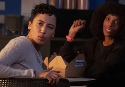 Duas mulheres em ambiente interno. Uma com cabelo encaracolado tem expressão séria. A outra, com afro, sorri e mostra o punho.