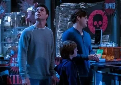 Dois homens e um menino observam em ambiente sombrio e azulado. Letreiros como Trophies com caveira e objetos peculiares no fundo.