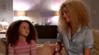 Mulher loira de pijama sorri para menina de cabelo cacheado em sofá. Ambas têm expressões felizes em um lar aconchegante.