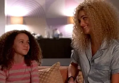 Mulher loira de pijama sorri para menina de cabelo cacheado em sofá. Ambas têm expressões felizes em um lar aconchegante.
