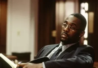 Homem negro de terno e óculos, com expressão séria, sentado em sala formal, segurando caderno e caneta.