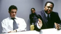 Dois homens em uma sala: um de terno gesticula intensamente, o outro, de camisa e gravata, olha pensativo. Uma mulher observa ao fundo.