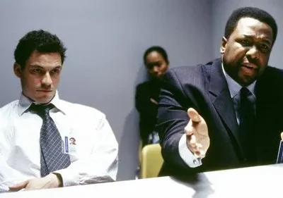 Dois homens em uma sala: um de terno gesticula intensamente, o outro, de camisa e gravata, olha pensativo. Uma mulher observa ao fundo.