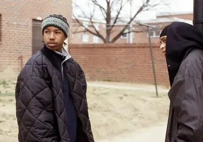 Dois jovens em roupas de inverno, um de gorro e jaqueta, outro de capuz, observam atentos em frente a prédios de tijolos.