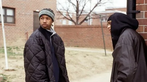 Dois jovens em roupas de inverno,um de gorro e jaqueta,outro de capuz,observam atentos em frente a prédios de tijolos.