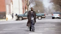 Homem negro sério de sobretudo e jeans, carregando arma visível, caminha por rua urbana. Cenário com carros e edifícios, clima tenso.