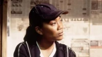 Jovem mulher de boné escuro e blusa esportiva olha pensativa para a direita, contra parede de jornais. Clima urbano sério.