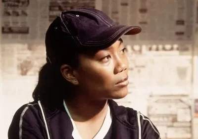 Jovem mulher de boné escuro e blusa esportiva olha pensativa para a direita, contra parede de jornais. Clima urbano sério.