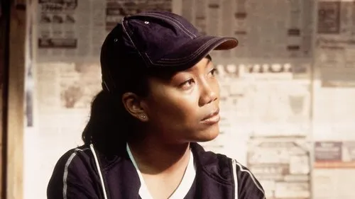 A Escuta – T01E08:Lições 1 Jovem mulher de boné escuro e blusa esportiva olha pensativa para a direita,contra parede de jornais. Clima urbano sério.