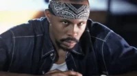 Homem com bandana preta e jaqueta jeans, olhar intenso e direto. Clima sério e urbano.