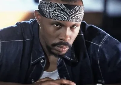 Homem com bandana preta e jaqueta jeans, olhar intenso e direto. Clima sério e urbano.
