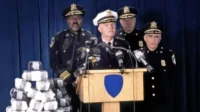 Coletiva de imprensa policial. Um oficial fala no púlpito, ladeado por outros e uma pilha enorme de drogas apreendidas. Clima sério.