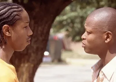 Dois jovens negros se encaram em close-up, conversando seriamente ao ar livre com um tronco de árvore no fundo.