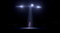 Noite escura. Nave triangular com luzes brilhantes irradia um feixe sobre figura de chapéu. Cena de suspense e mistério.