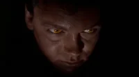 Rosto de homem parcialmente iluminado, com olhos brilhantes amarelo-dourados que se destacam na escuridão. Clima sombrio e tenso.
