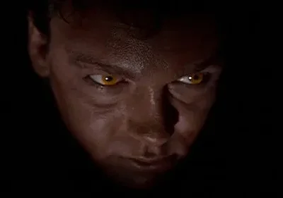 Rosto de homem parcialmente iluminado, com olhos brilhantes amarelo-dourados que se destacam na escuridão. Clima sombrio e tenso.