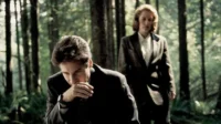 Homem de terno na floresta escura, mão no rosto. Mulher séria de blazer atrás. Atmosfera misteriosa, investigativa.