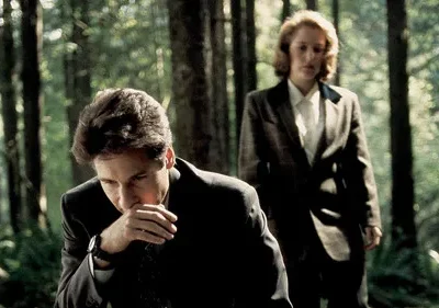 Homem de terno na floresta escura, mão no rosto. Mulher séria de blazer atrás. Atmosfera misteriosa, investigativa.