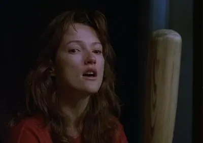 Mulher com cabelo bagunçado e olhar assustado, marca no nariz. Um taco de beisebol ao lado. Cenário escuro e tenso.
