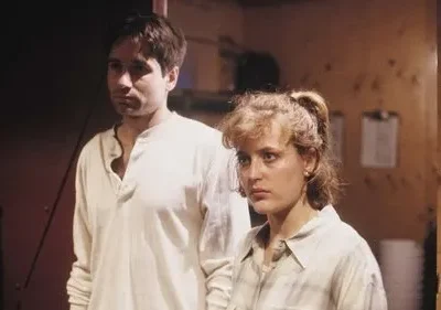 Um homem e uma mulher, ambos sérios, olham para direções diferentes. Estão num ambiente interno rústico e mal iluminado.