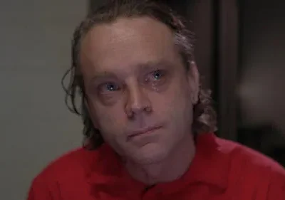 Homem de meia-idade com cabelo desarrumado, camisa vermelha e lágrimas nos olhos, expressa tristeza num fundo escuro.