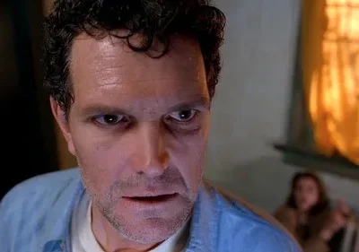 Homem com olhar intenso no primeiro plano. Ao fundo, mulher assustada e desfocada em um ambiente escuro e tenso.
