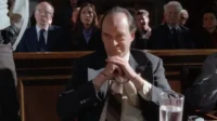 Um homem de terno, algemado, com olhar desafiador em um tribunal. Pessoas assistem ao fundo. Cena de drama legal.