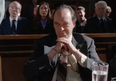 Um homem de terno, algemado, com olhar desafiador em um tribunal. Pessoas assistem ao fundo. Cena de drama legal.