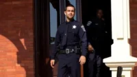 Banshee – T01E01:Piloto 9 Um policial sorridente em uniforme azul-marinho sai de um edifício de tijolos. Outro policial sombrio está na porta atrás dele.