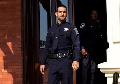 Um policial sorridente em uniforme azul-marinho sai de um edifício de tijolos. Outro policial sombrio está na porta atrás dele.