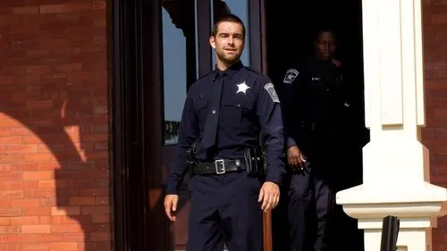 Um policial sorridente em uniforme azul-marinho sai de um edifício de tijolos. Outro policial sombrio está na porta atrás dele.